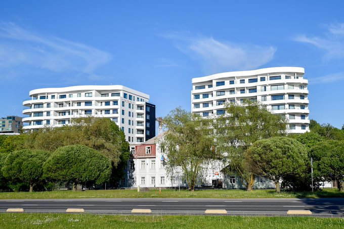 Two modern, curved white apartment buildings stand behind a row of green trees and a small white house with a red roof under a blue sky.