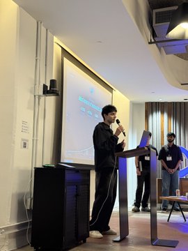 A speaker stands at a podium with a laptop, addressing an audience while a presentation is projected on the screen behind him, with a few attendees visible in the background. The image captures a talk at what appears to be a Monad Foundation event, reflecting the post's appreciation for the foundation's support of teams building on the Monad blockchain.