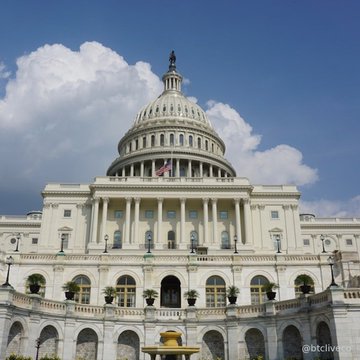 A photograph of the United States Capitol building featuring its iconic white dome against a bright blue sky with scattered clouds, with a watermark reading 'btclive' in the bottom right.
