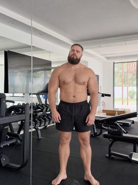 A shirtless, bearded man wearing dark athletic shorts standing barefoot in what looks like a home gym with exercise equipment and a large TV in the background.