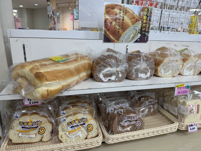 A display of various packaged Japanese breads including rolls and chocolate-filled pastries, with a promotional photo card showing a cream roll and price tags like ¥2300 and ¥1200 visible.