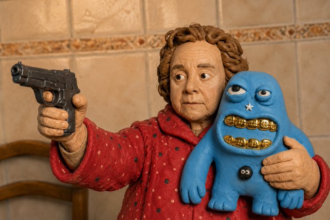 A stop-motion claymation figure of an elderly woman in a red polka-dot robe holds a black handgun in one hand while clutching a blue, toothy monster figurine with gold teeth in the other, set against a tiled wall background.