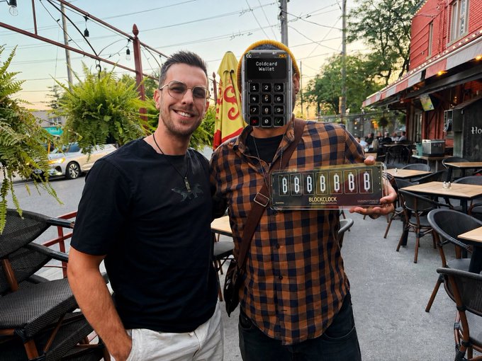 Two men posing on a street; one wears a black shirt and the other holds a digital card wallet device displaying '6BBB6' and a Blockclock showing '6666666'.