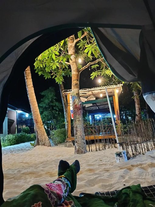 A first-person view from inside a dark tent looking out at sandy ground, trees, and a warmly lit wooden structure at night.