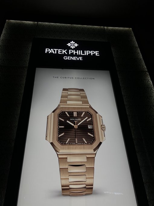 A Patek Philippe advertisement display showcasing a rose gold Cubitus collection watch with a brown dial, date window, and integrated bracelet against a black background.