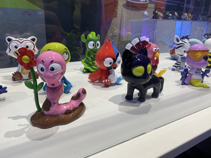A display of colorful vinyl/designer toy figures in a glass case, including a pink worm-like creature holding a flower, a green figure, a red character, and a black spiked creature.