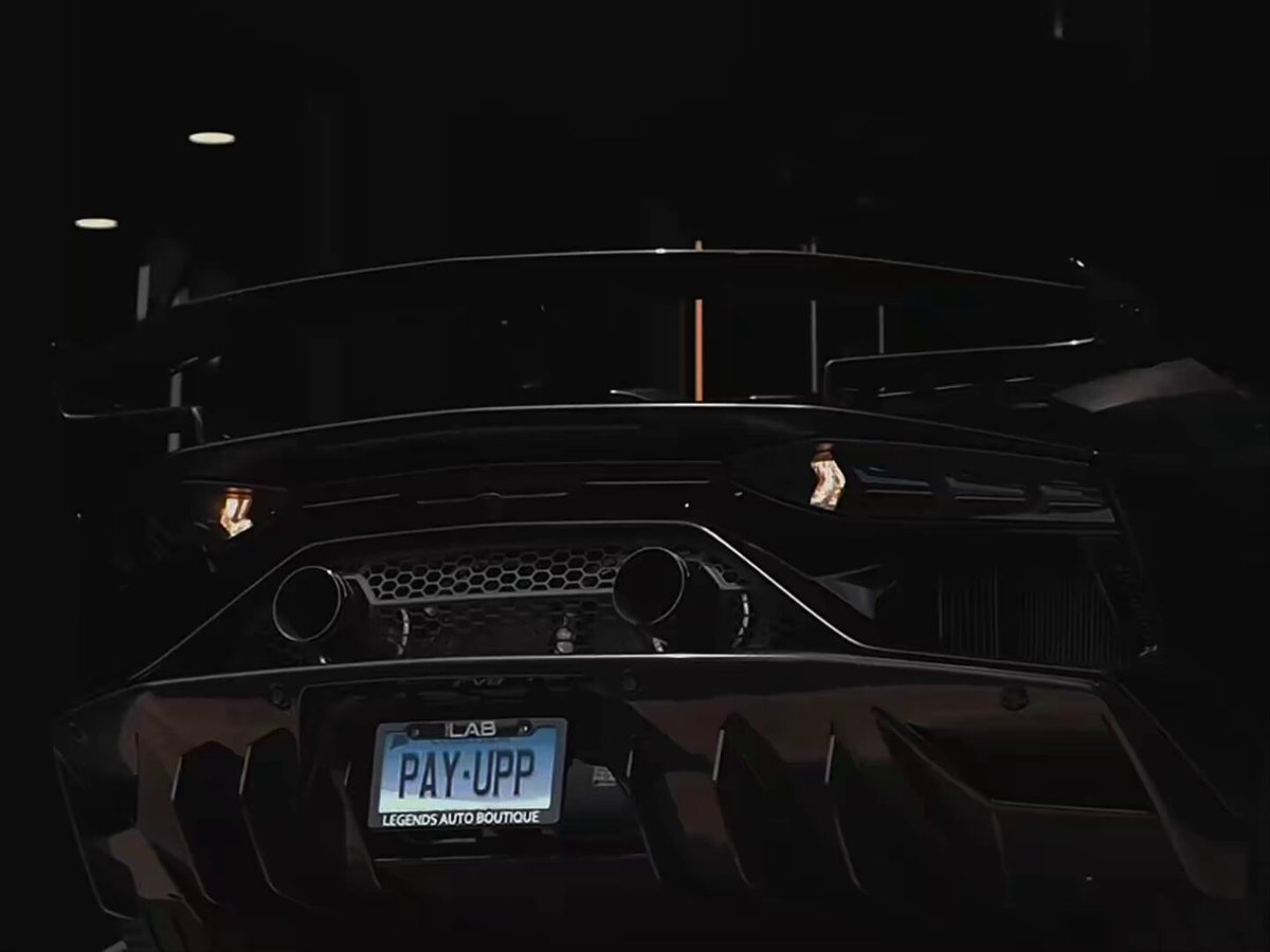 The image shows a dark-colored sports car with a license plate that reads "POV". It appears to be parked indoors, possibly at a garage or showroom. The car's rear end features two exhaust pipes, adding to its sporty appearance. The background is dark and out of focus, drawing attention to the car in the foreground. The image also contains text that reads "POV: your rich life when you start trading weather markets on Polymarket...", which seems to be a promotional message related to the sports car shown in the photo.