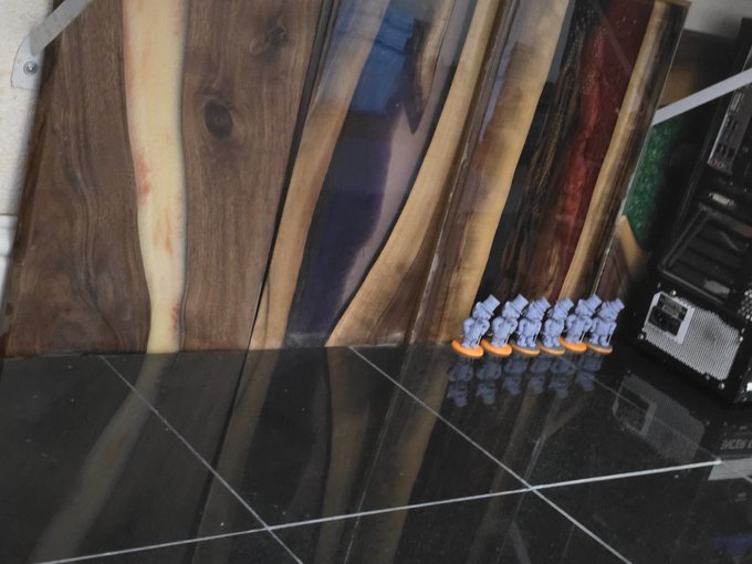 A display of handcrafted wooden boards featuring various wood grains and epoxy resin inlays, with a row of small painted miniature figures lined up on the black tiled floor.