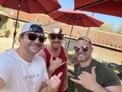 Two men wearing sunglasses, hats, and casual shirts pose outdoors under red umbrellas, smiling and gesturing towards the camera.