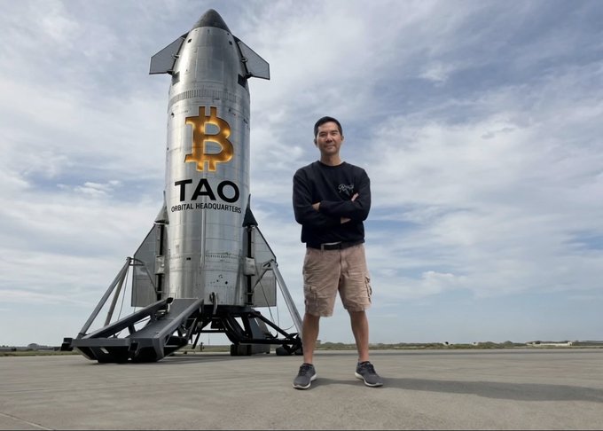 A man stands with his arms crossed in front of a large rocket decorated with a Bitcoin symbol and the text 'TAO DIGITAL HEADQUARTERS' on a tarmac.