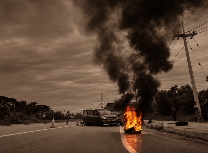 A dramatic sepia-toned photograph of a car on fire with thick black smoke billowing into a cloudy sky on a road with a power line visible.