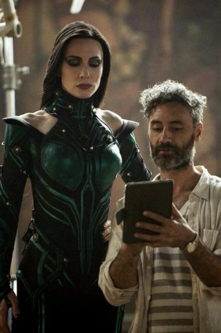 A woman in a green and black ornate costume stands next to a bearded man who is looking at a tablet device, appearing to be a behind-the-scenes moment from a film or photo shoot.