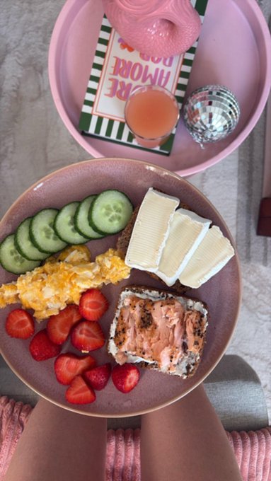 A pink tray holds a book titled "More Good" alongside a pink beverage, while a larger plate displays a balanced brunch spread of scrambled eggs, cucumber slices, brie cheese, salmon-topped toast, and fresh strawberries—perfectly illustrating the post's reminder to "eat well, get outside, and touch some grass."
