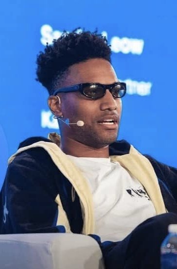 A man wearing dark sunglasses and a beige hoodie over a white t-shirt speaks into a headset microphone while seated on a white couch at the "SALT New York" conference, with partial text "K..." visible on the blue backdrop behind him. The image relates to the accompanying post featuring Ansem, who is known for commenting on cryptocurrency tokens like $LAPTOP that experienced massive losses after insiders profited.