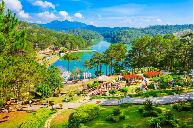 An aerial view of a lush green park with a lake, garden areas, and mountains in the background under a blue sky.