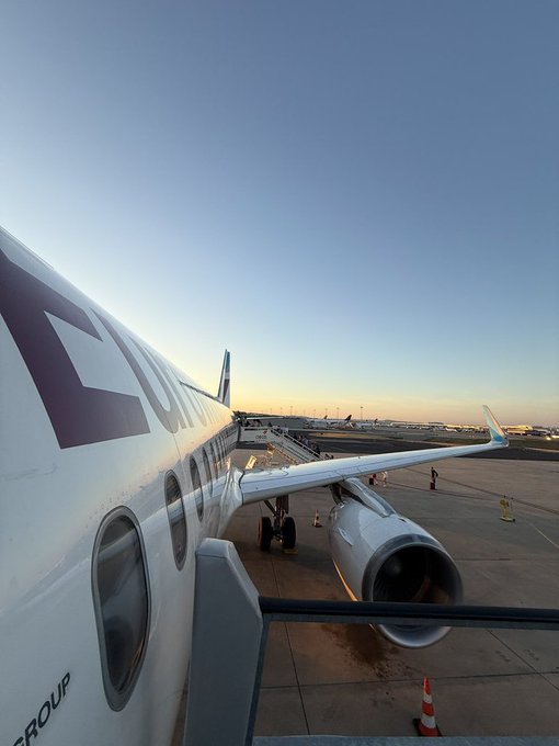 View from a passenger window showing the wing and engine of an airplane on the tarmac at sunset, with the text 'GROUP' partially visible on the fuselage.