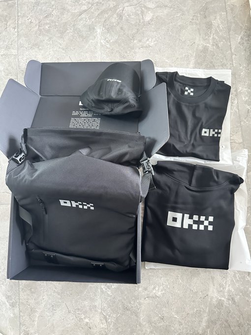 An open black box containing OKX-branded merchandise, including a black backpack, t-shirt, and cap, all featuring the 'OKX' logo.