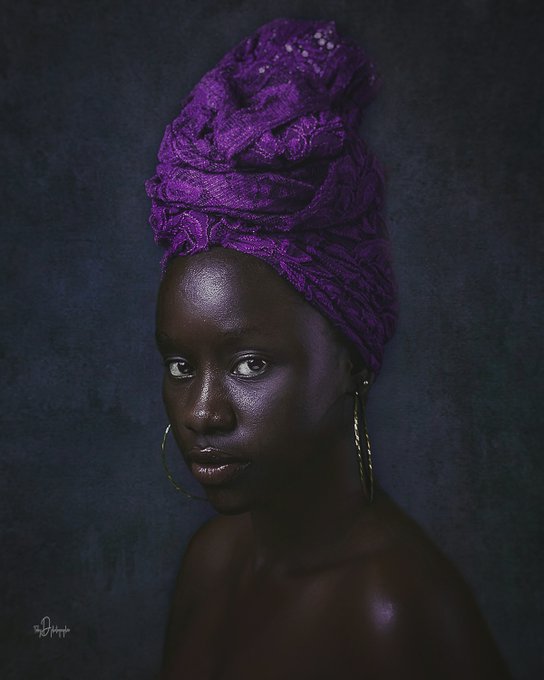 A striking portrait of a woman wearing a vibrant purple head wrap and gold hoop earrings, photographed against a dark blue textured background.