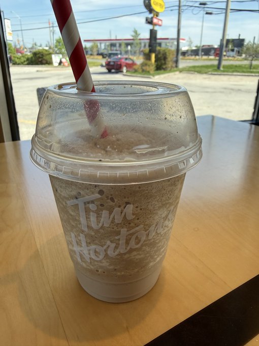 The image features a Tim Hortons ice cream drink sitting on top of a wooden table with a straw sticking out of its lid. The background shows a street view, indicating that this scene takes place outdoors or near an urban setting. There are also cars visible in the distance, suggesting that it is located in a busy area.