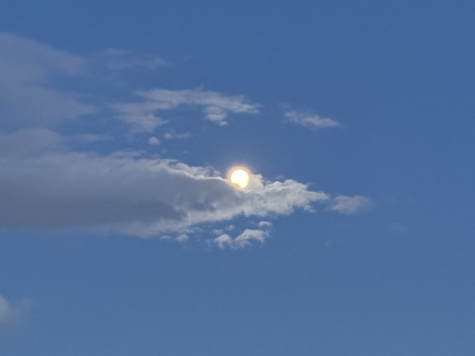 The sun shining brightly through a thin layer of clouds in a clear blue sky.