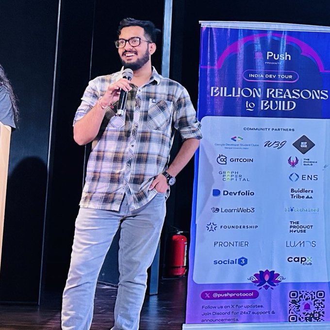 A bearded man wearing glasses and a plaid shirt speaks into a microphone beside a banner reading 'Push INDIA DEV TOUR - BILLION REASONS to BUILD' listing various community partners including Gitcoin and Devfolio.