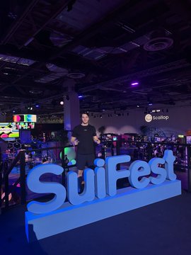 A man poses behind a glowing blue "SuiFest" sign at the Sui Network's conference, embodying the lively atmosphere and strong community presence highlighted in the post about the event's great vibes.