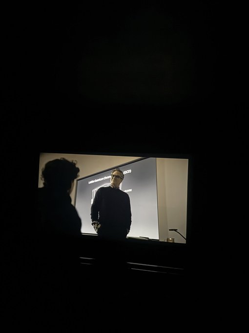 A silhouette of a person sits in a darkened auditorium watching a presenter on a screen displaying "London Business School" text. The scene captures the quiet anticipation of arriving early before an event begins.