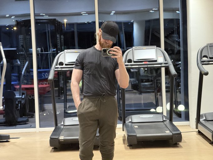 A man wearing a black cap, black t-shirt, and olive green joggers takes a mirror selfie in a gym, standing in front of treadmills.