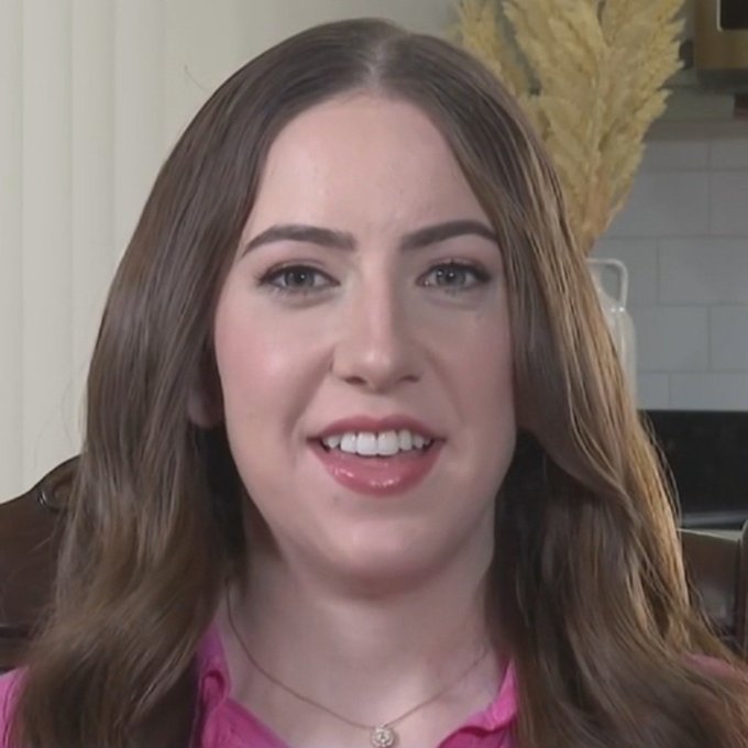 A close-up photo of a woman with long brown hair, wearing a pink top and a delicate pendant necklace, smiling at the camera.