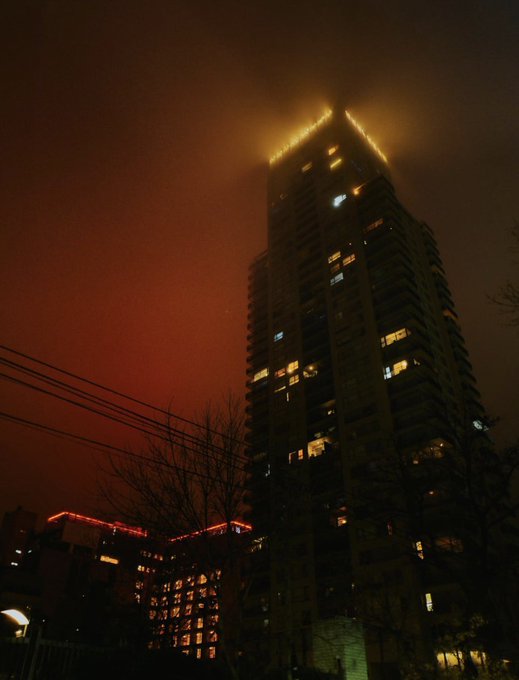 A nighttime photograph of a tall illuminated apartment tower against a red-orange sky, with power lines and bare trees in the foreground.