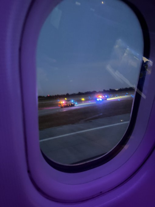 A view through an airplane window at dusk shows emergency vehicles with flashing red, blue, and amber lights on the tarmac, suggesting the plane may have been involved in an incident. The caption "Oh great" conveys the passenger's frustration or resignation at the situation.