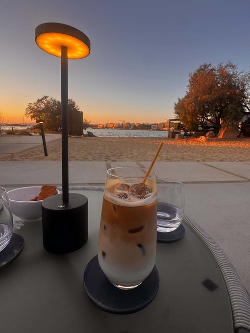 An iced coffee drink in a tall glass sits on an outdoor table beside a glowing orange table lamp, with a scenic waterfront sunset and city skyline in the background.