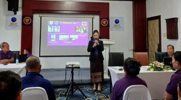 A woman stands speaking into a microphone at an indoor presentation, with a projector screen displaying images behind her and an audience seated in front.