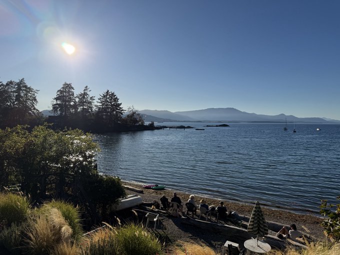 A scenic waterfront view at golden hour shows people sitting on a rocky shore with a small boat, calm water, distant mountains, and sailboats under a bright sun.