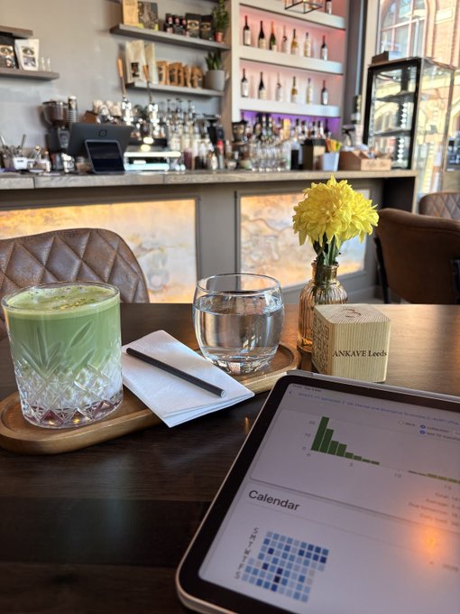 A cozy café scene showing a wooden table with a green matcha latte, a glass of water, a yellow chrysanthemum in a vase, and a tablet displaying a calendar app, with a coffee bar visible in the background.