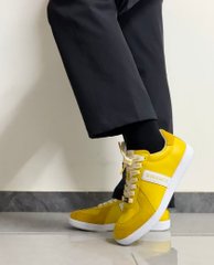 A close-up shot focusing on a person's lower legs and feet, showing them wearing dark pants and bright yellow sneakers with white soles and laces, standing on a grey floor.