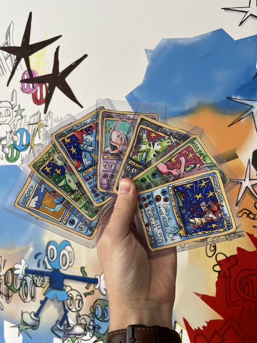 A hand holds a fanned-out spread of illustrated Pokémon trading cards against a colorful background featuring doodle art and star patterns.