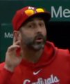 A man with a beard wearing a red "Can" (likely Canada) cap and red hoodie gestures with his hand while speaking, with a partial "Me" visible in the background.