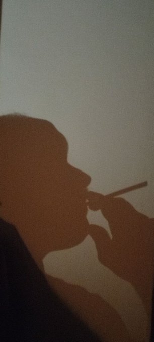 A dramatic silhouette shadow of a person wearing a cap and smoking what appears to be a cigarette against a light-colored wall.