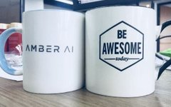 Two white coffee mugs side by side; the left one reads "AMBER AI" and the right displays "BE AWESOME today" inside a hexagon.