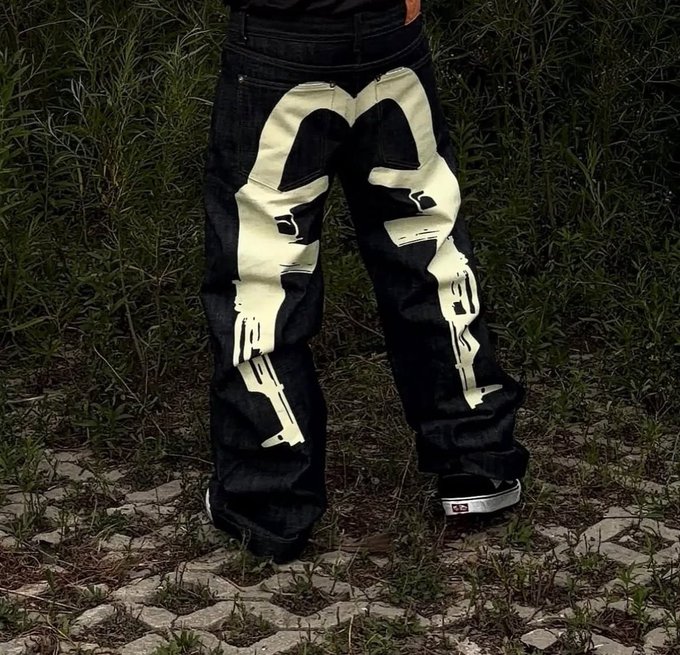 A close-up shot of black denim jeans featuring large white painted skull face designs, paired with black sneakers, standing on a paved surface with grass in the background.