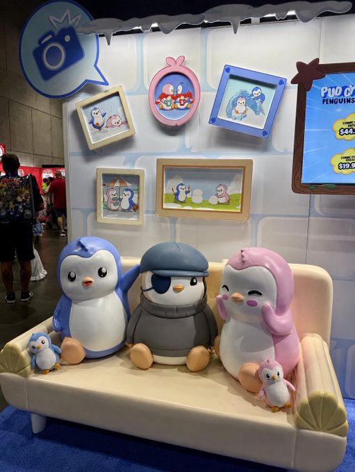 A display at a convention or store featuring three large plush penguin toys (blue, grey with an eyepatch, and pink) sitting on a small beige couch, with framed penguin artwork on the wall behind them and a 'Pudgy Penguins' screen visible to the right.
