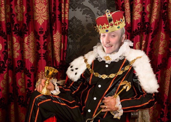 A photo of a man dressed as a king wearing an ornate red and gold crown, fur-trimmed royal robe, and gold chain, holding a goblet against a backdrop of red and gold patterned curtains.