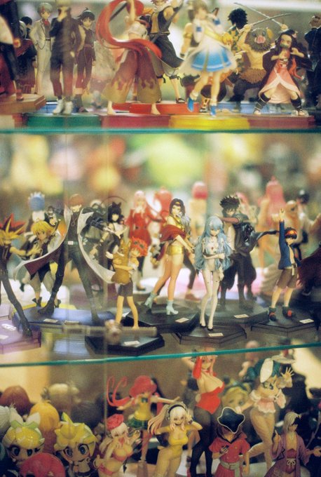 A glass display case is filled with shelves of colorful anime figurines from various series, featuring characters in dynamic poses.
