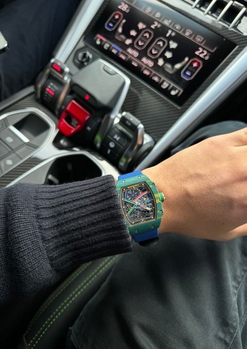 A wrist wearing a blue-strapped skeleton watch with a tonneau-shaped case is shown resting on a denim-clad leg, with a luxury car's center console and digital climate controls visible in the background.