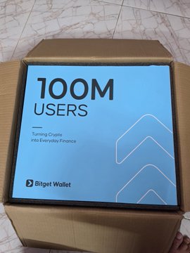 Photograph of an opened cardboard box containing promotional materials for Bitget Wallet celebrating '100M USERS' with the tagline 'Turning Crypto into Everyday Finance.'