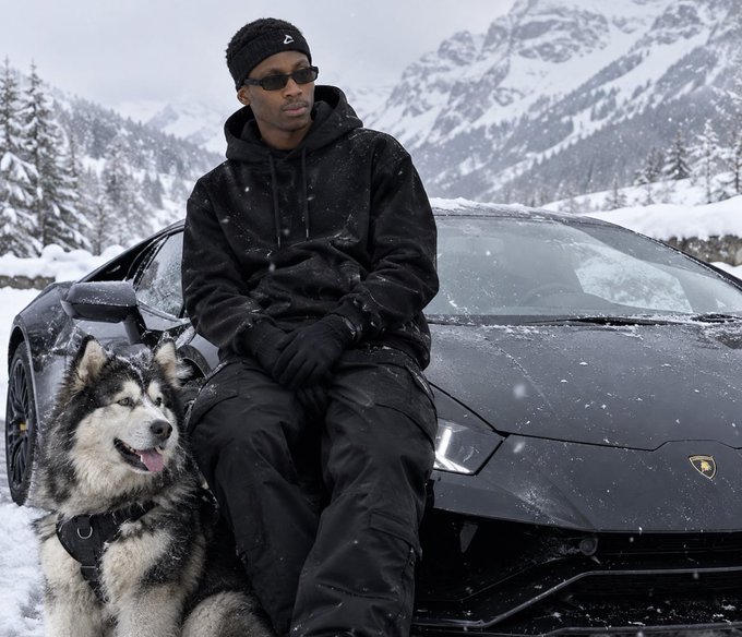 A man in all-black attire with a beanie and sunglasses sits on the hood of a black Lamborghini in a snowy mountain setting, accompanied by a large husky dog wearing a tactical harness.