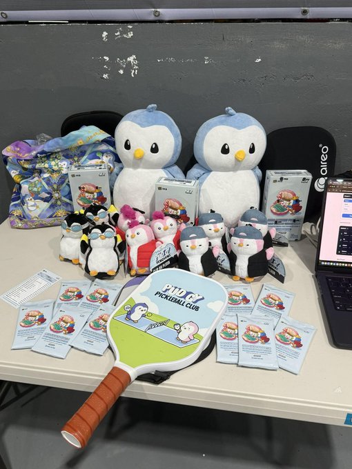 A display table featuring JTT Pickleball Club merchandise, including penguin plush toys, small penguin figurines, a pickleball paddle, and promotional cards arranged around the merchandise.