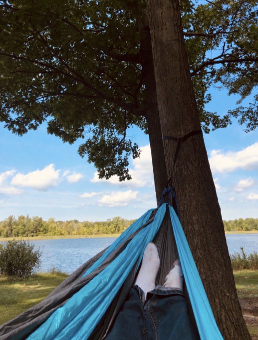 A blue hammock hangs from a tree by a lake, with a person's bare feet visible inside it.