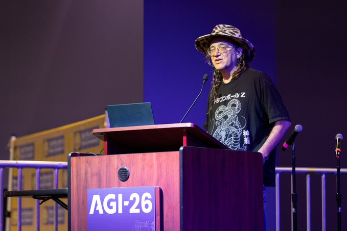 A person wearing glasses, a patterned hat, and a black Japanese t-shirt speaks at a podium labeled 'AGI-26' during a presentation with a laptop and microphone.